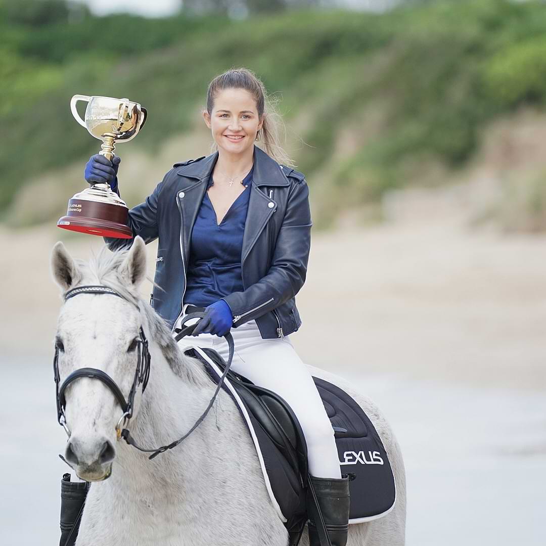 Jockey Michelle Payne sits on a white horse with a black Lexus saddle. She is holding the 2021 Melbourne Cup trophy in her right hand. They are on the beach.