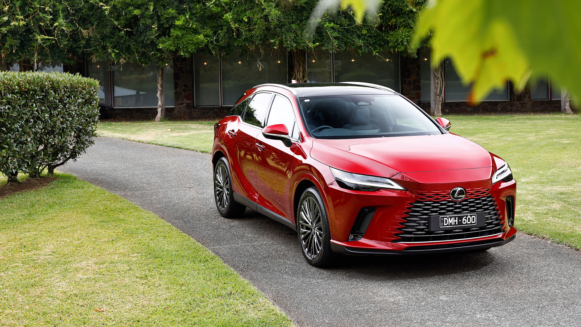 Red Lexus RX 450h+ plug-in hybrid luxury SUV parked on a paved driveway next to a grassy area.