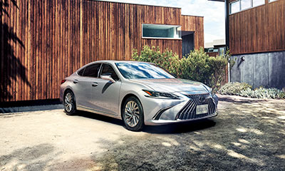 Silver Lexus ES compact crossover parked at an angle in front of a modern, wooden house.