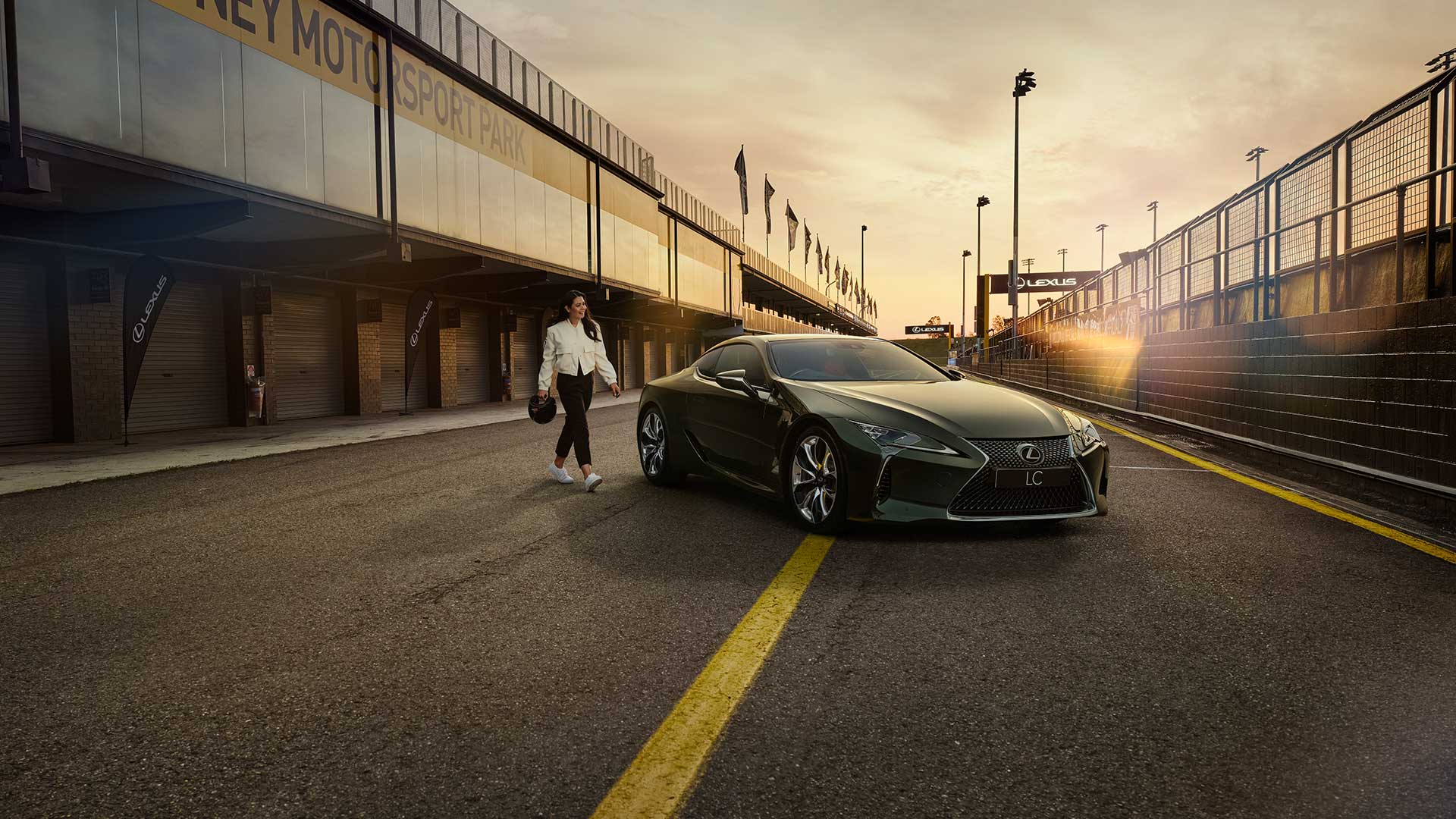 A woman dressed in a white jacket and black pants and holding a helmet walks towards an LC coupe parked in a pit lane